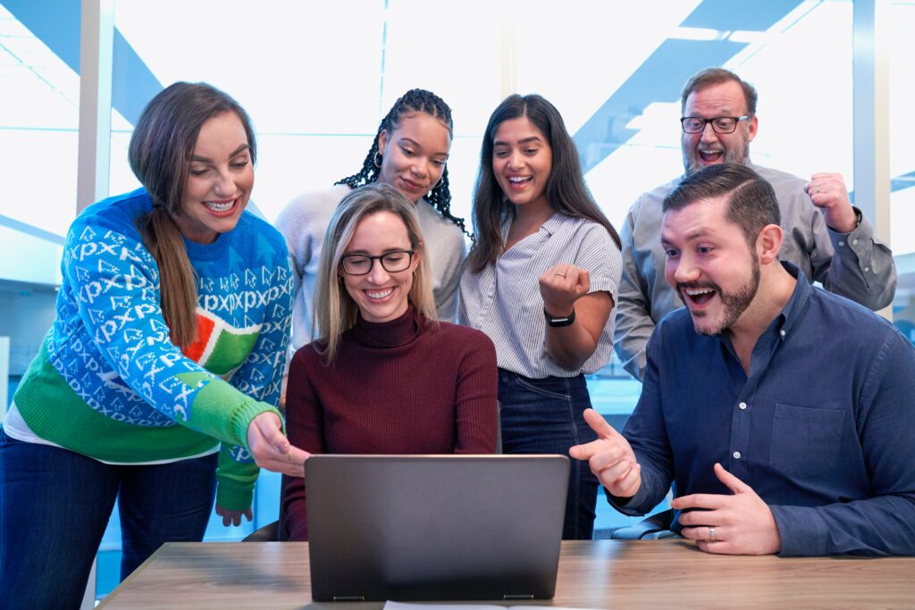 6 people looking at one laptop laughing and pointing 