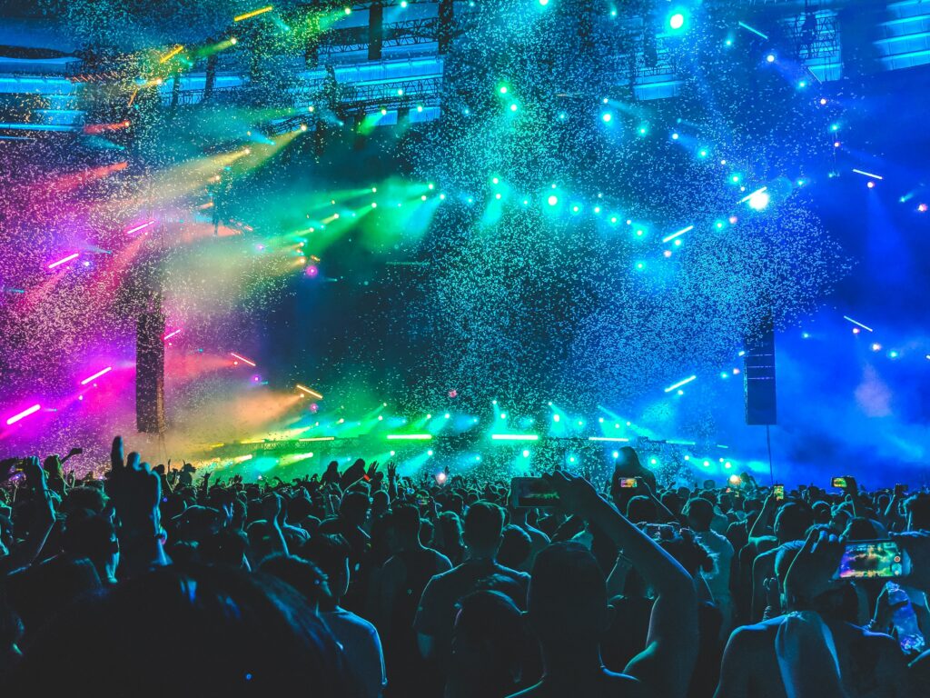 A group of people partying at the club in the dark with colourful lights on the ceiling and the stage area. 