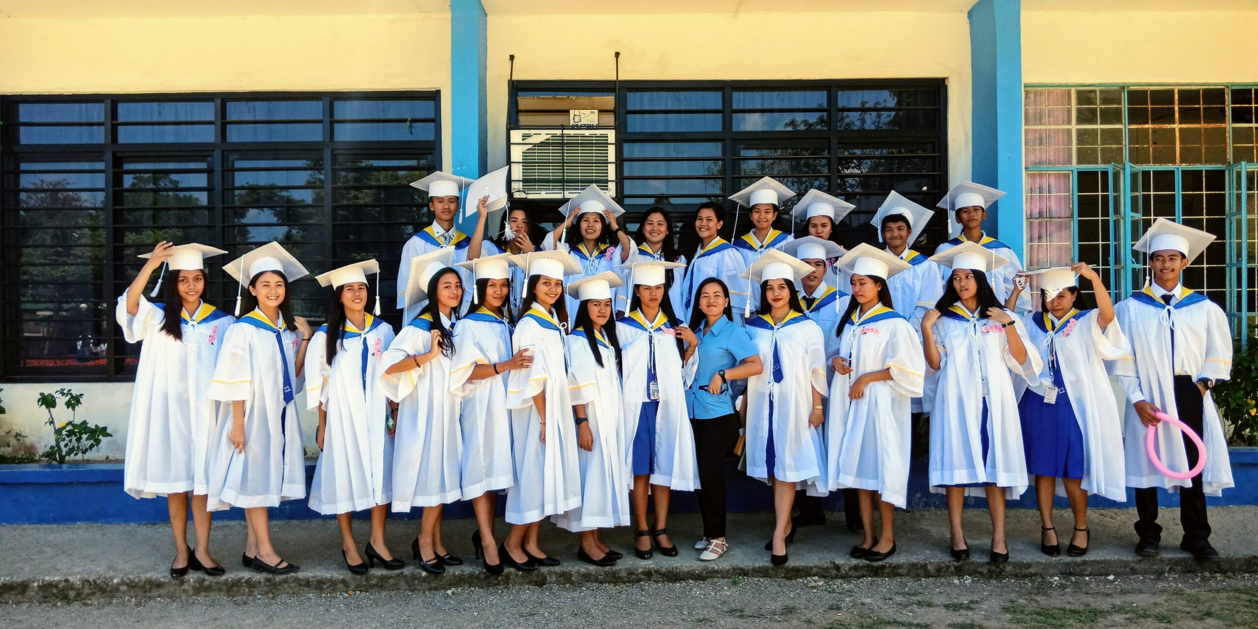 A group of girls graduating wearing all white and with their graduate hat. Their teacher is also in the middle of the group wearing blue.