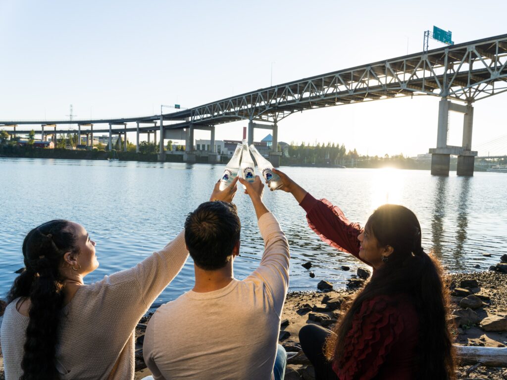 Three friends sitting on the sand whilst looking at the view of a bridge. 