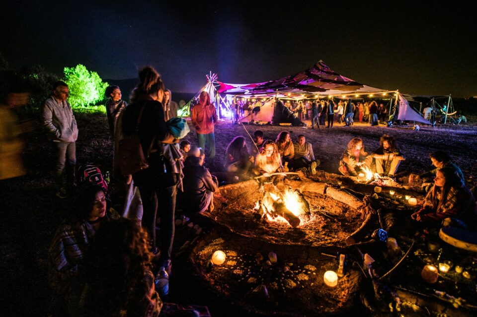 A group of people sitting around a fire with a tent behind them which has vibrant colours