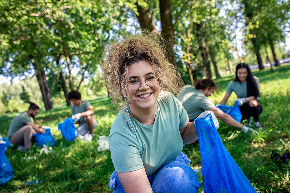 Portrait of volunteering young woman with garbage bag cleaning city park with friends.