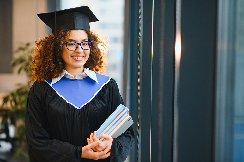 Female student smiling, wearing a graduation cap and gown, holding books, celebrating academic achievement and future success