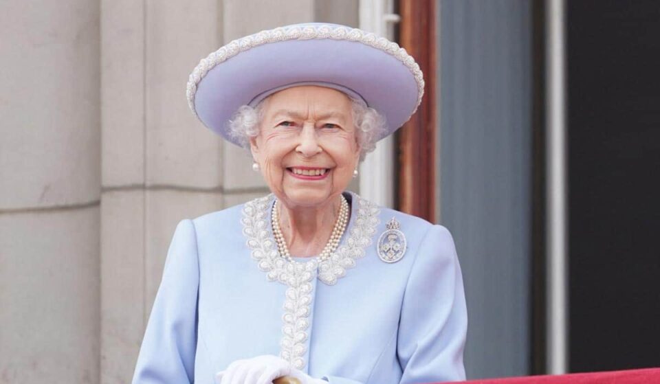 A picture of Queen Elizabeth II on the balcony in a blue suit jacket and matching hat with pearls