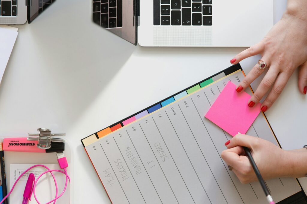 a person writing on a pink sticky note with a laptop next to them
