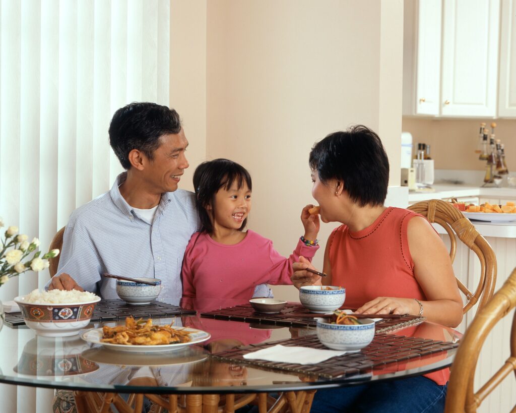 A little girl feeding her mum food whilst on her dad's lap 