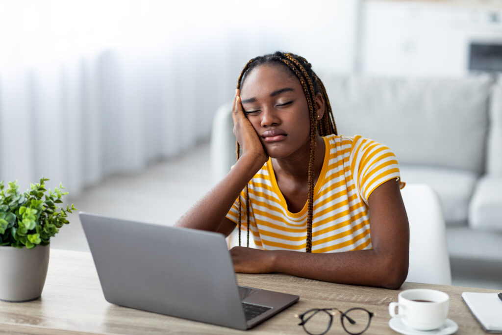 Young black woman sitting at table, leaning on hand, sleeping in front of laptop. 