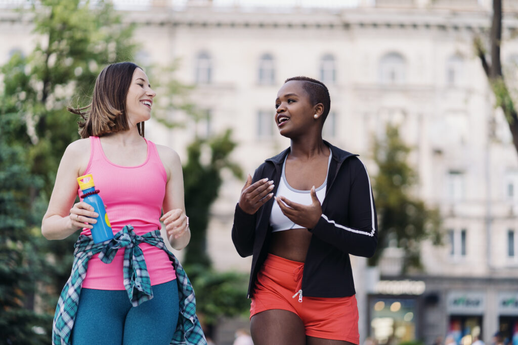 Cheerful smiling friends in sportswear walking after a sport session in the city discussing. Multiethnic women having a fitness workout break.