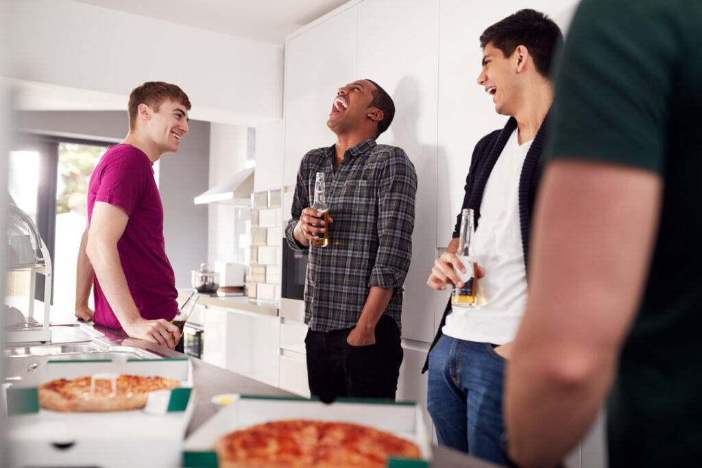 Group of university students in shared house kitchen drinking and eating pizza together.