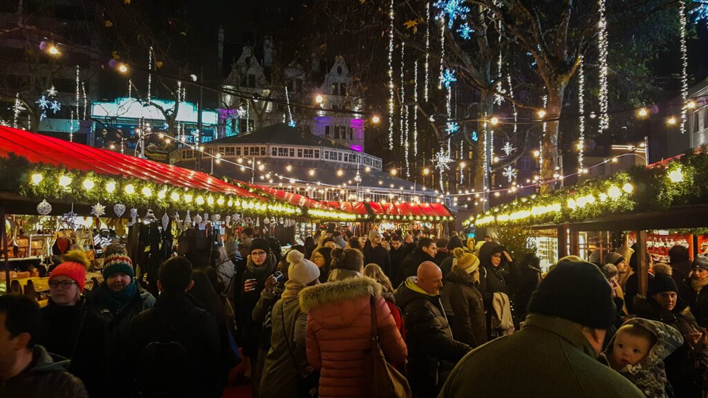People walking through a Christmas market with stalls next to them. There are also Christmas light hanging from the stalls 