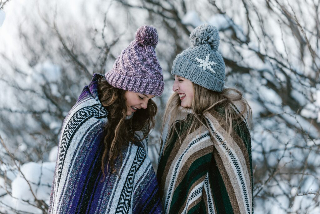 Two friends laughing in the snow with blankets around them 