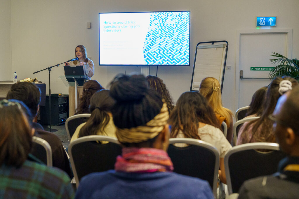 A woman standing and talking into a mic whilst there is a group listening.