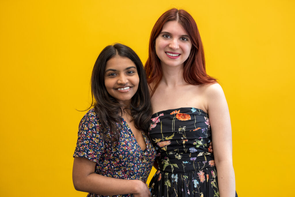 Two girls standing next to each other smiling behind a yellow background.