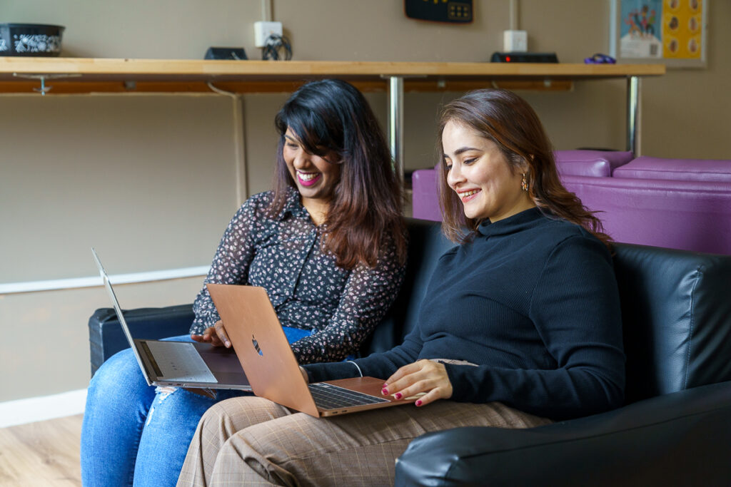 Two girls sitting on a sofa with there laptops open.