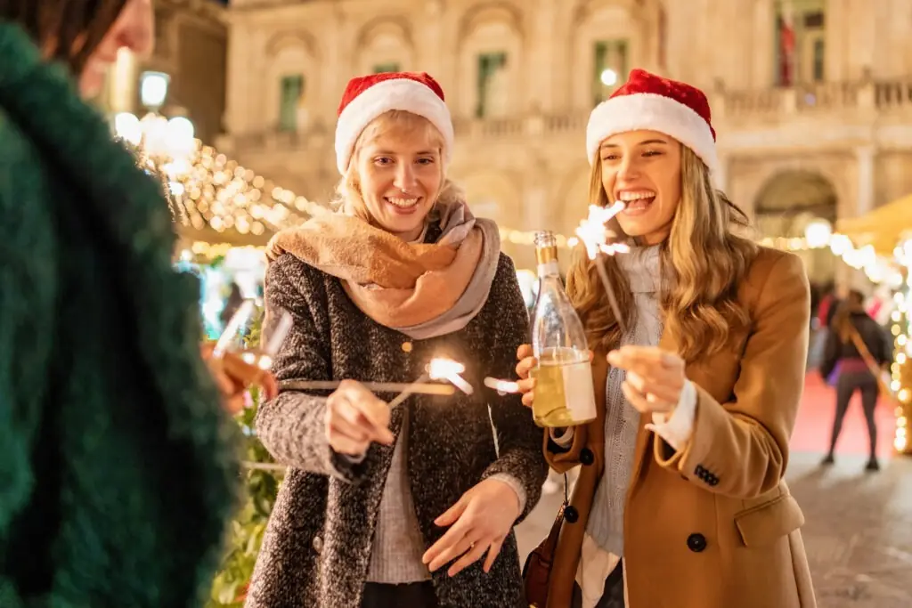 Cheerful young women in Santa hats celebrating at a festive Christmas market. Image captures joy of holiday season with sparkling lights, warm drinks, and historic architecture backdrop.