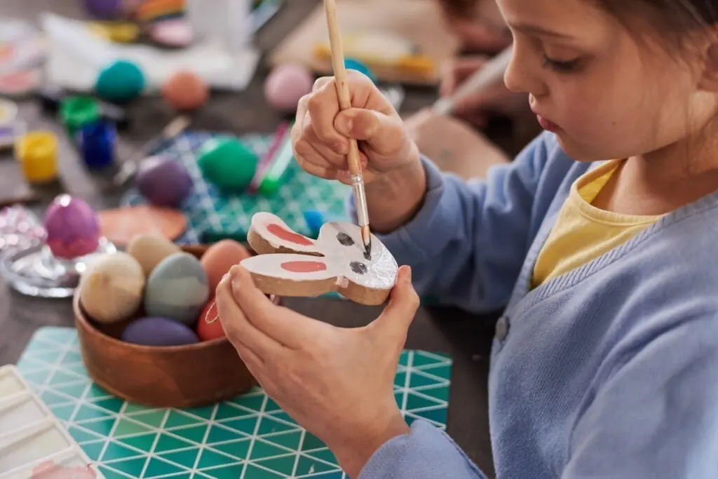 Close-up of little girl sitting at the table making and painting figure of rabbit for Easter holiday