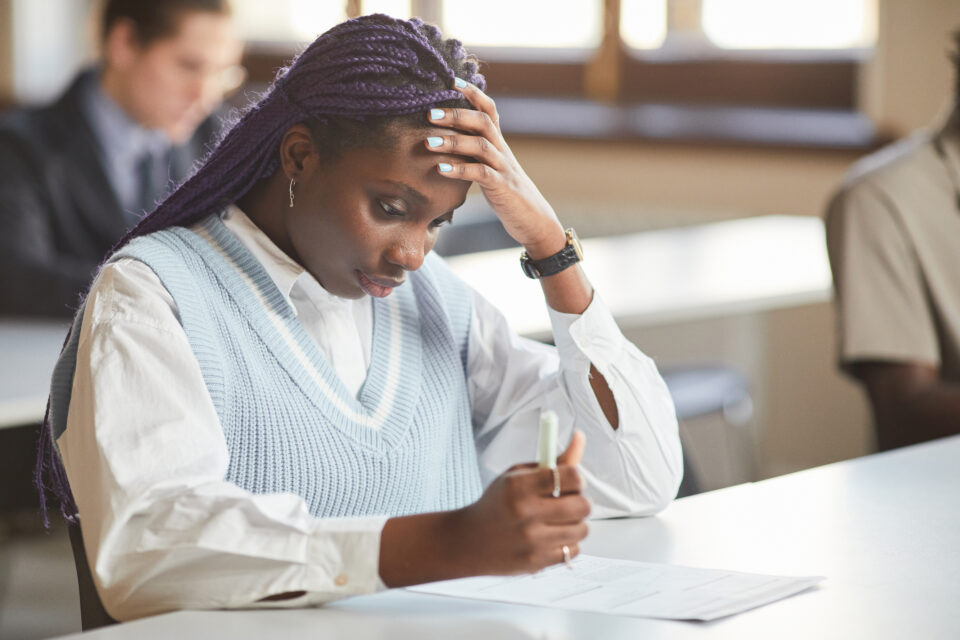 Portrait of young African-American woman taking exam in school while sitting at desk in college and thinking, copy space