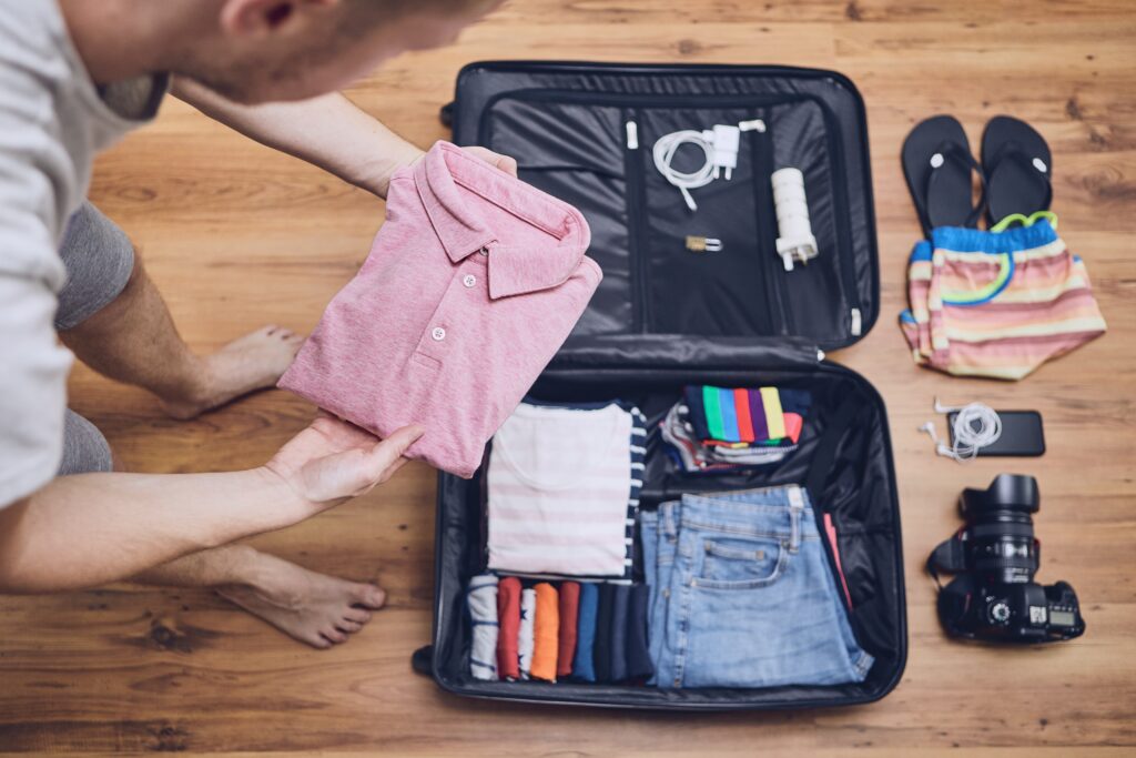 Preparing for trip. Young man packing clothing for vacation, flip flop, camera and other things on hardwood floor.