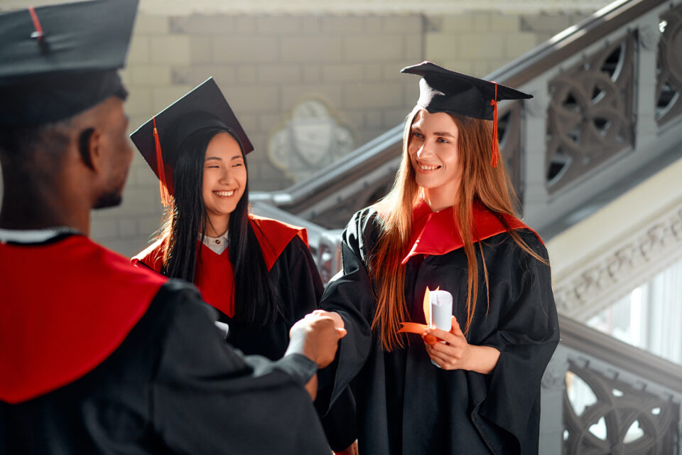 Diverse group of happy students in graduation gowns and caps celebrate academic achievements with smiles at joyful ceremony, bonding and sharing congratulations during milestone success