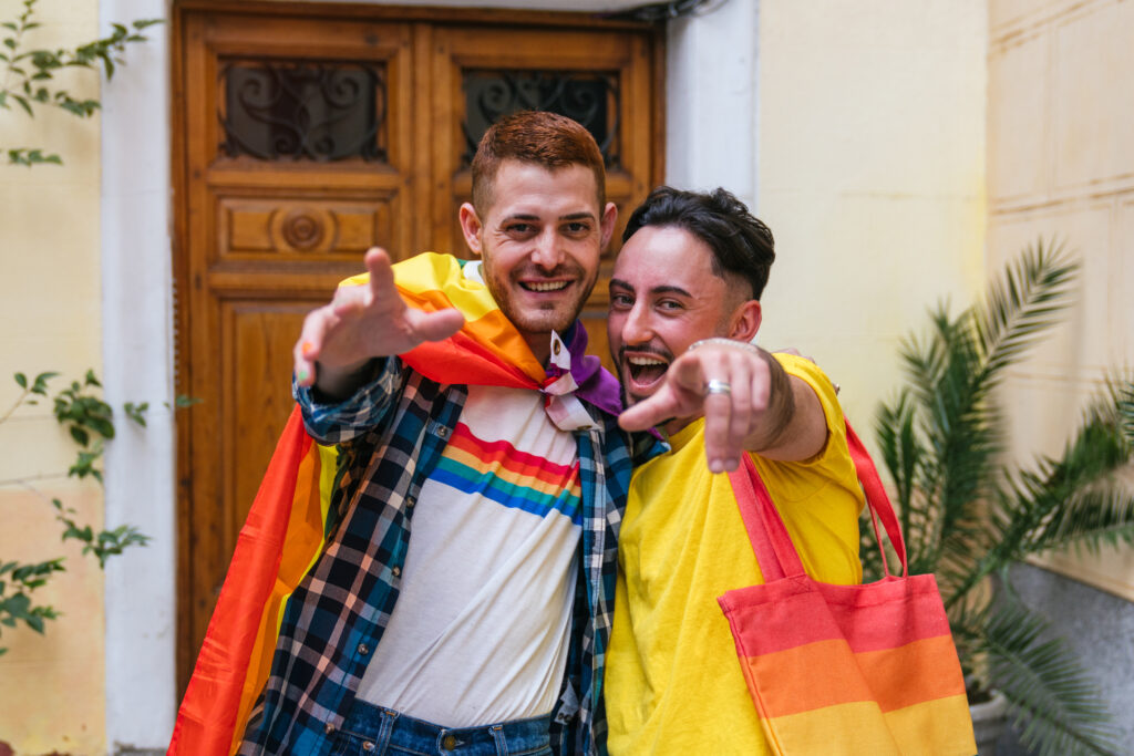 Two gay friends exuding happiness and pride, embracing tightly, pointing joyfully outside their adorned doorway, showcasing LGBT symbols.