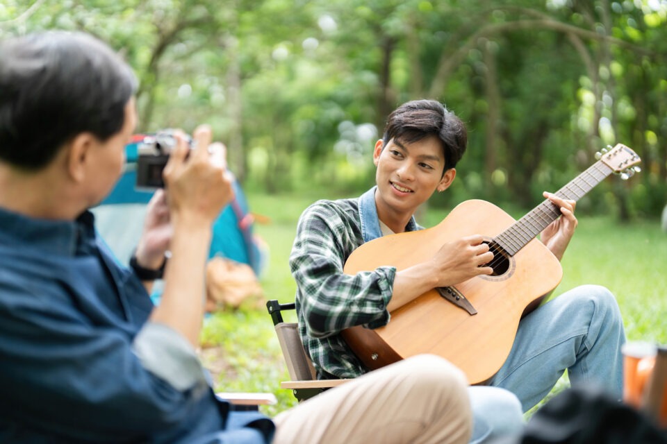A delightful camping scene where a father photographs his son playing guitar, emphasizing family connection and outdoor fun.